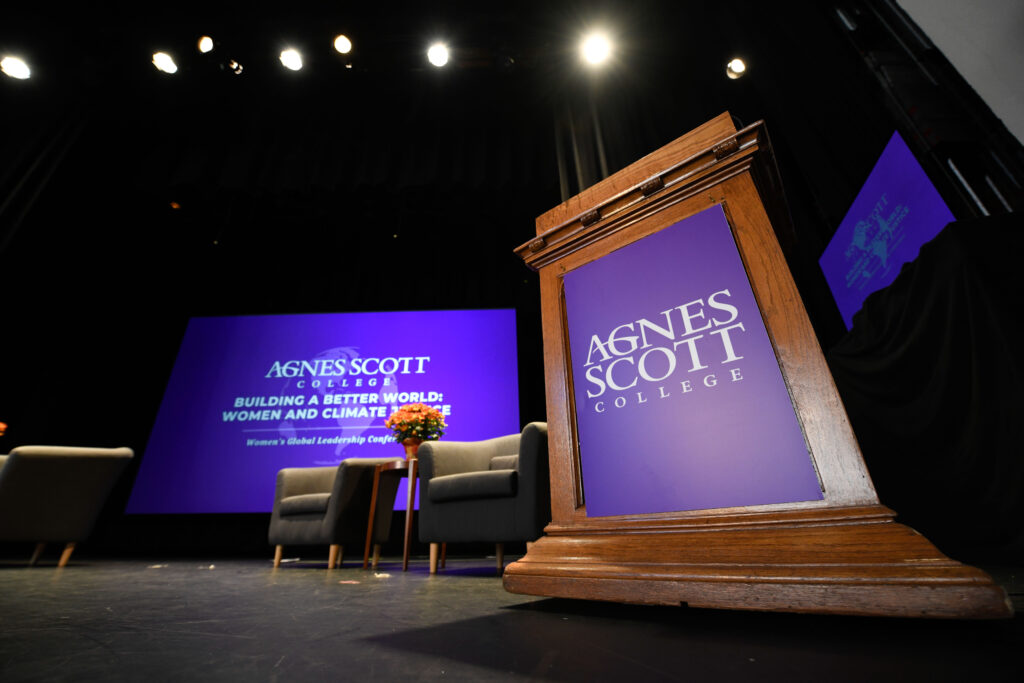 A podium sits on a stage with a purple banner that reads "Agnes Scott College." Behind the podium are chairs placed in an interview style and a projection of a presentation slide sits in the back with the words: Agnes Scott College Building a Better World: Women and Climate Justice Women's Global Leadership Conference